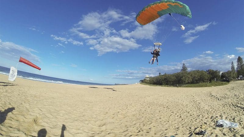 Experience the thrill of tandem skydiving at Sunshine Coast Beach, landing on golden sand with a vibrant green and orange parachute.