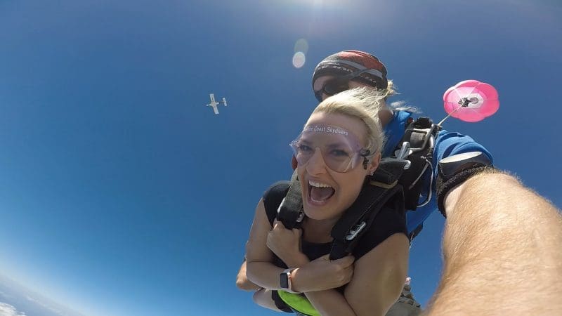 Woman smiling with excitement during Sunshine Coast Beach Tandem Skydive, aeroplane overhead, parachute open against clear blue sky.
