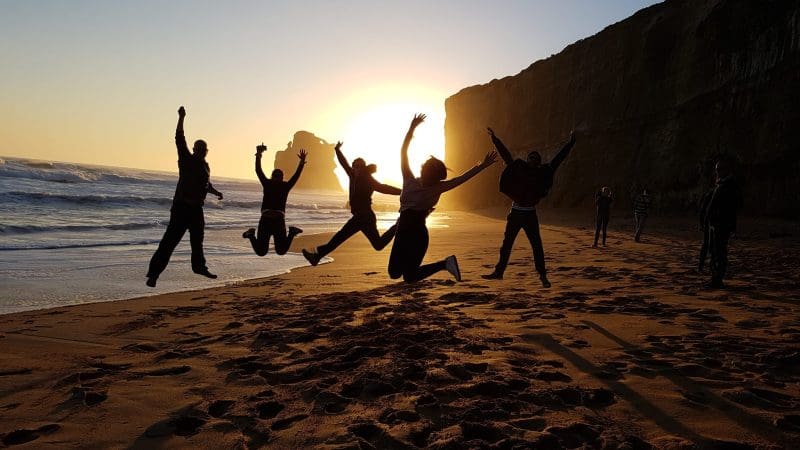 Energetic group silhouettes jumping on a sandy beach at sunset during Go West Tours’ 1 Day Great Ocean Road Sunset Tour, Australia.