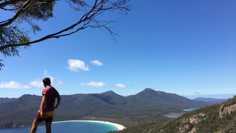Explorer on a rocky ledge overlooks Wineglass Bay Tour, Richmond, forests, and mountains under bright clear skies—Tasmania adventure.