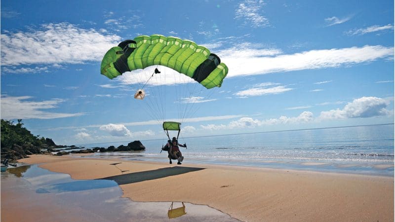Skydiver with a green parachute from Cairns Tandem Skydive Extras lands on pristine sandy beach by turquoise sea waters.