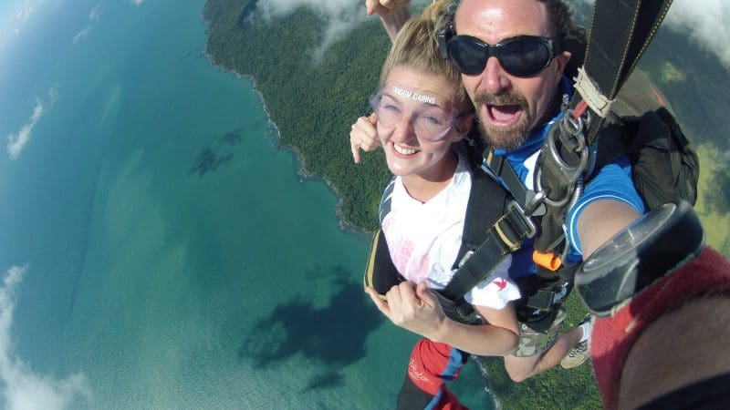 Experience the thrill of Cairns tandem skydiving—two adventurers soaring over stunning coastline, grinning above ocean and rainforest.