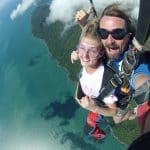 Tandem skydivers free-fall above scenic coastline, grinning at camera, with blue ocean waves and lush green landscape below.