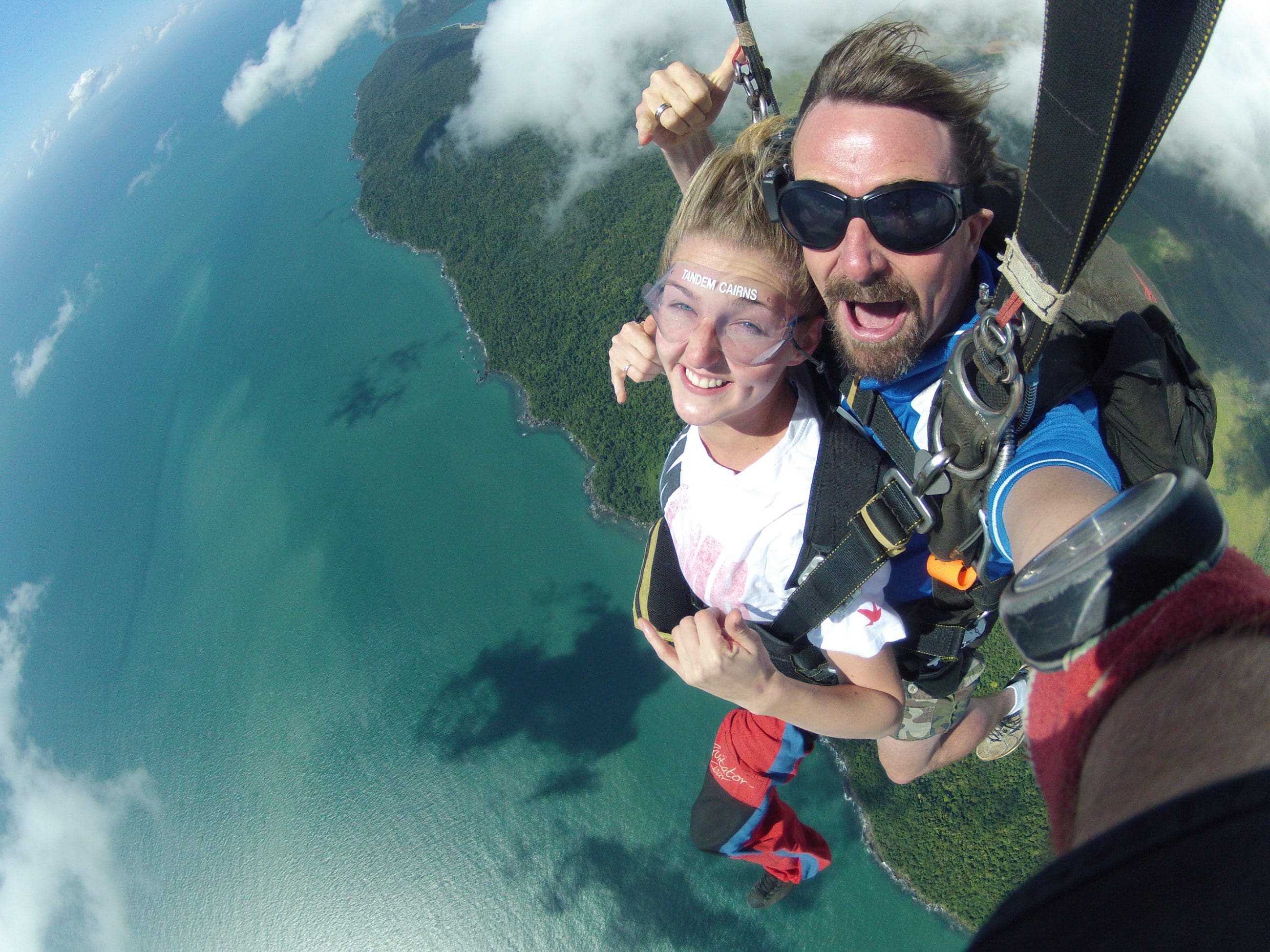 Tandem skydivers free-fall above scenic coastline, grinning at camera, with blue ocean waves and lush green landscape below.