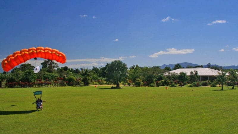 Skydiver descends with vibrant orange parachute onto lush grassy field after thrilling Cairns Tandem Skydive Extras adventure.