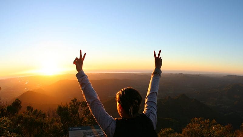 Person triumphantly raising arms at sunrise atop mountain after Hinterland Magic Glow Worm Experience, celebrating scenic adventure.