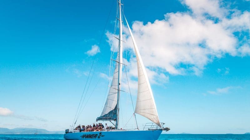 Hammer 2 Day 2 Night Whitsunday Sailing group cruises crystal-clear blue waters under a vibrant sky dotted with scattered white clouds.