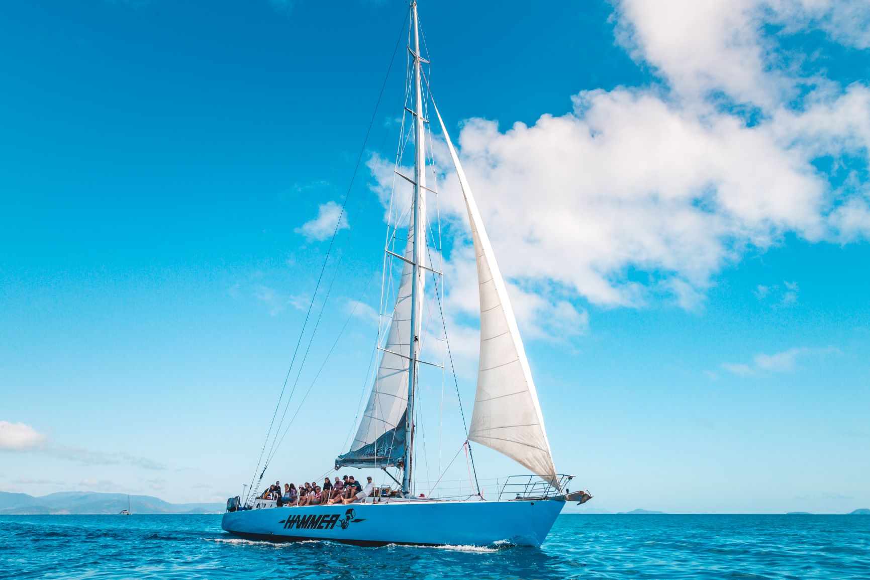 Hammer 2 Day 2 Night Whitsunday Sailing group cruises crystal-clear blue waters under a vibrant sky dotted with scattered white clouds.