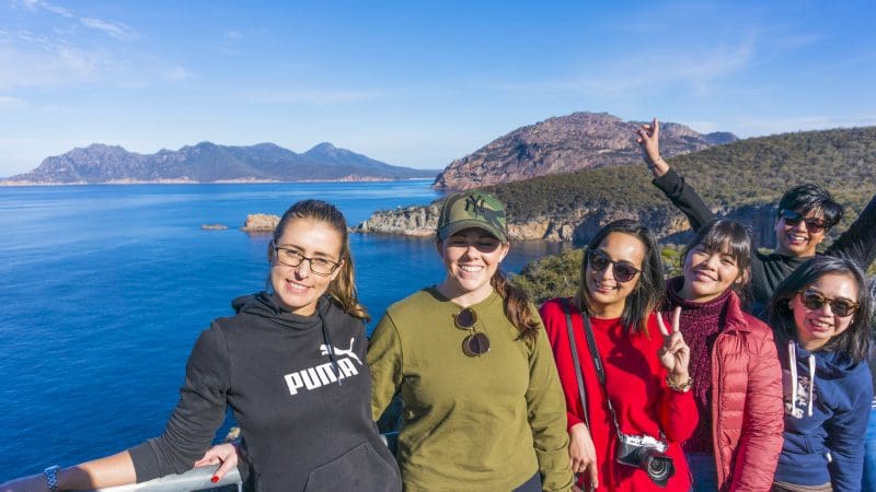 Six friends smiling by a scenic railing overlooking blue water and mountains on a Wineglass Bay Full Day Tour via Richmond, Tasmania.