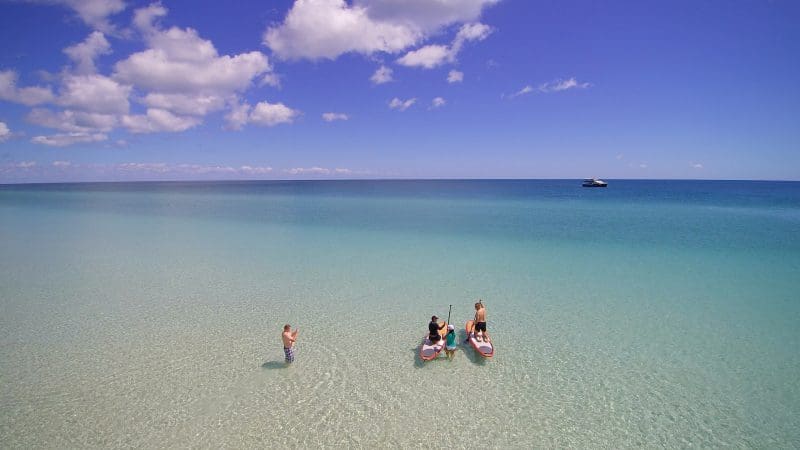 Tourists paddleboarding in crystal-clear, shallow waters during the Remote 1 Day K'gari Fraser Island tour under a blue, clouded sky.