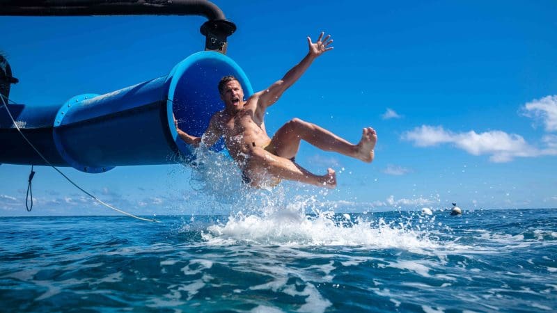 Man plunges into the crystal-clear sea from a vibrant blue waterslide at Outer Reef Fitzroy Island under a sunny, cloudless sky.