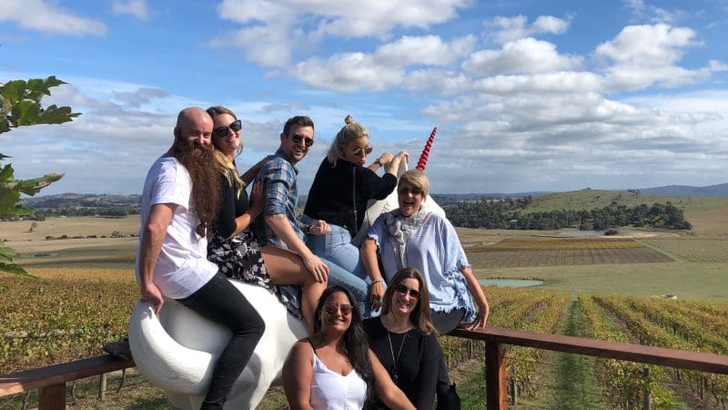 Six friends pose atop a giant white unicorn at a picturesque Yarra Valley winery, enjoying a relaxed, fun wine tour experience.