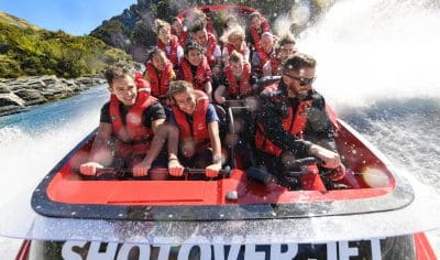 Tourists in red life jackets thrill on the Shotover Jet, smiling as water splashes during an adventure ride in Queenstown, New Zealand.
