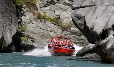 Experience the adrenaline rush as a red Shotover Jet boat races through Queenstown's rocky canyon with thrilled passengers on turquoise water.