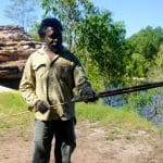 Experience an authentic Kakadu Arnhem Land Tour: a man outdoors demonstrates traditional spear skills amid lush trees and rugged rocks.