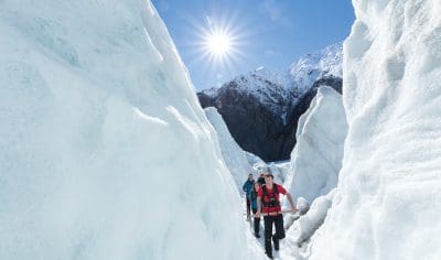 Three hikers trek a narrow icy glacier path under bright sun, surrounded by towering snowy mountains and breathtaking glacial views.