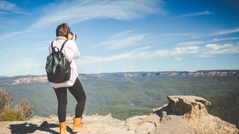 Adventurer with rucksack photographing panoramic Blue Mountains vista from rugged cliff edge on Uncharted Tour, sweeping valley below.
