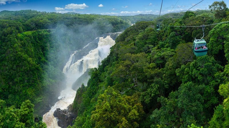 Experience a Skyrail cable car soaring above vibrant rainforest and a stunning waterfall beneath clear blue skies on the way to Rainforestation.