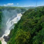 Skyrail cable car travels above vibrant rainforest canopy, overlooking a majestic waterfall under a bright blue sky with fluffy clouds.