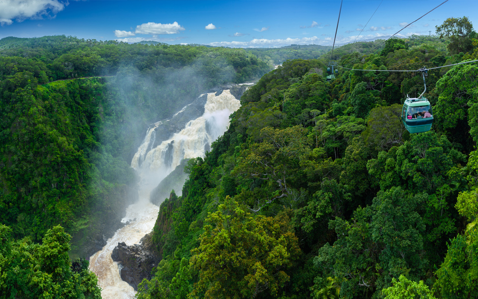 Skyrail cable car travels above vibrant rainforest canopy, overlooking a majestic waterfall under a bright blue sky with fluffy clouds.