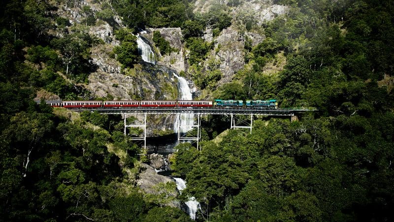 The iconic red and green Grand Kuranda train crosses a scenic bridge near a cascading waterfall, surrounded by lush rainforest.
