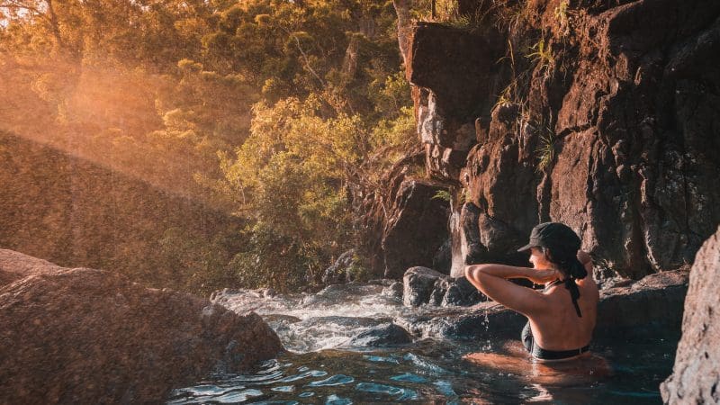 Swimmer unwinds in a crystal-clear rocky pool beneath Falls To Paradise Waterfall, with sunlight streaming through lush forest trees.