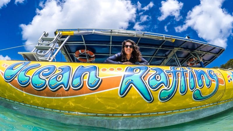 Happy woman leans over vibrant Ocean Rafting Northern Exposure Tour boat in crystal-clear water beneath a bright, blue sky with clouds.