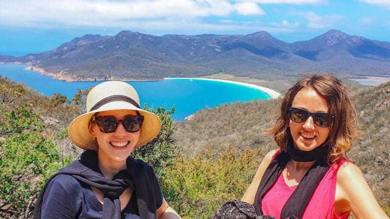 Two women in sunglasses smile by stunning Wineglass Bay on a full-day Richmond tour, with clear skies and breathtaking scenery.