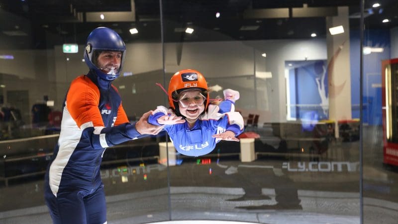 A child wearing professional skydiving kit floats with an instructor inside an iFLY indoor wind tunnel, enjoying realistic flight.