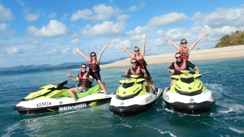 Group of five on two vibrant yellow jet skis, smiling and raising arms during Airlie Adventure Jetski Tour by a scenic sandy beach.