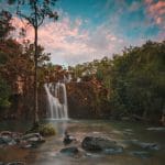 Falls To Paradise Waterfall cascades into a tranquil pool, framed by lush trees and rocks beneath a vibrant sunset, natural beauty.