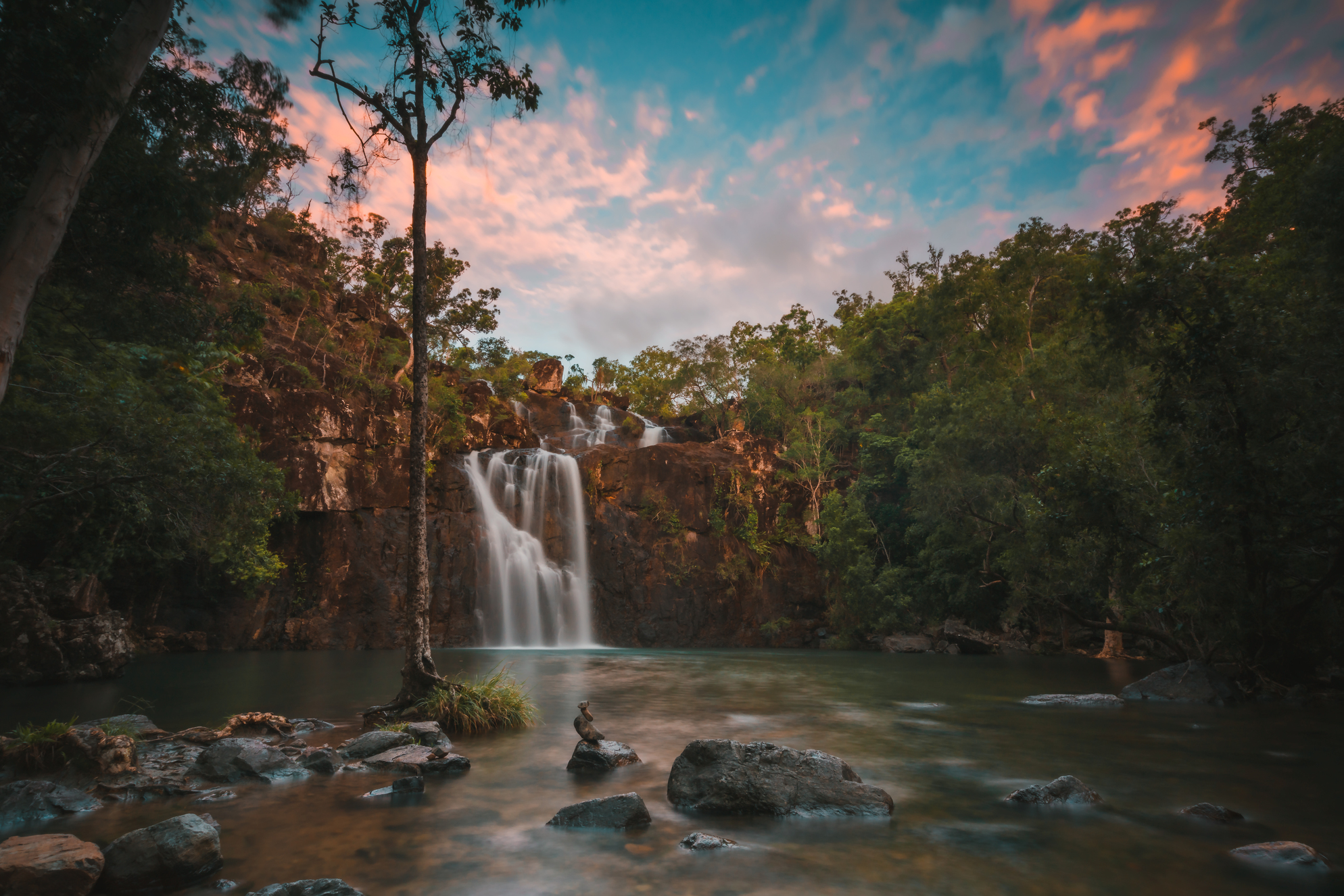Falls To Paradise Waterfall cascades into a tranquil pool, framed by lush trees and rocks beneath a vibrant sunset, natural beauty.