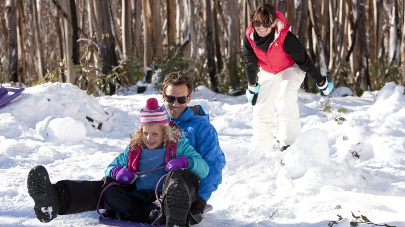 Family sledges down a snowy Lake Mountain hill during their 1 Day Melbourne Snow Waterfalls Tour, smiling in a winter wonderland.