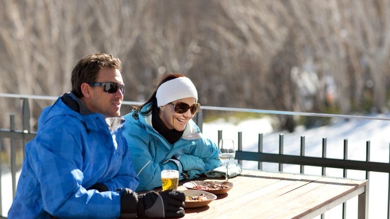 Two travellers in winter jackets savour snacks and drinks after a Lake Mountain Snow Waterfalls Tour near Melbourne, enjoying the scenery.