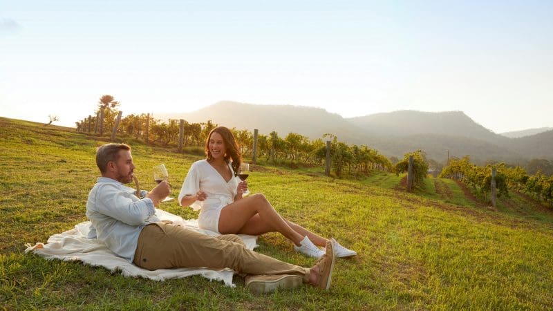 A couple enjoys a scenic sunset picnic on a blanket in Hunter Valley’s vineyard during an exclusive 1 Day Wine Tour experience.