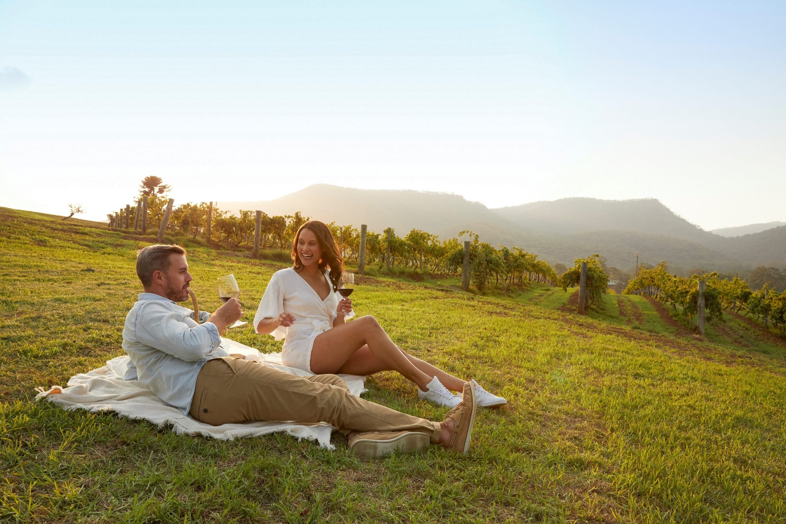 Couple enjoying premium wine on a scenic Hunter Valley tour, relaxing on a blanket with sweeping mountain views at sunset.