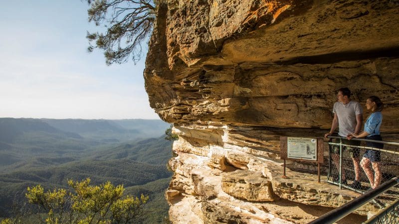 Two travellers at a Blue Mountains lookout enjoy panoramic sunset views of lush forested valleys on a 1 Day Wilderness Tour.