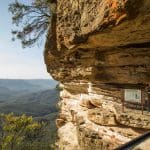 Two tourists admire a breathtaking sunset over Blue Mountains wilderness, overlooking lush forest valleys and dramatic mountain scenery.