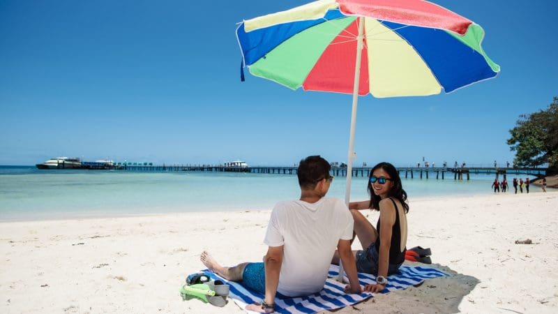 Couple relaxing on striped towel beneath vibrant umbrella at Green Island Half Day 9AM Departure, enjoying tropical paradise views.