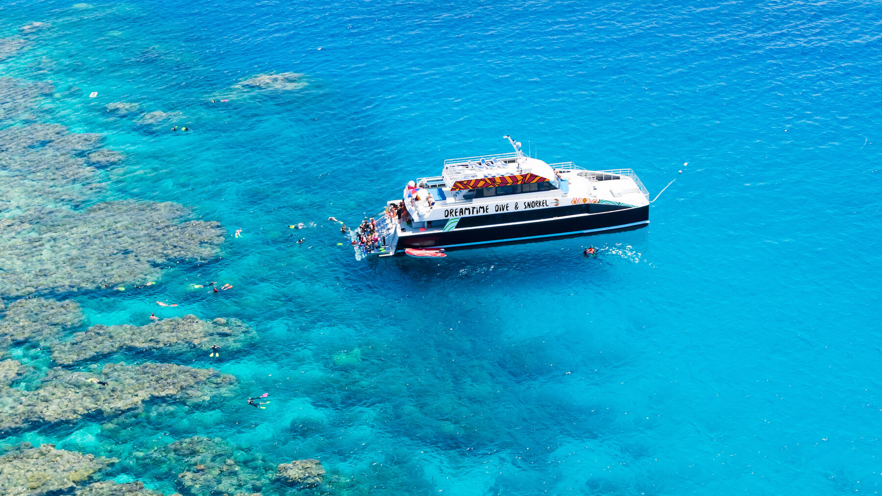 A boat anchored in crystal-clear blue water by a vibrant reef, with guests experiencing a full-day guided snorkel and dive adventure.