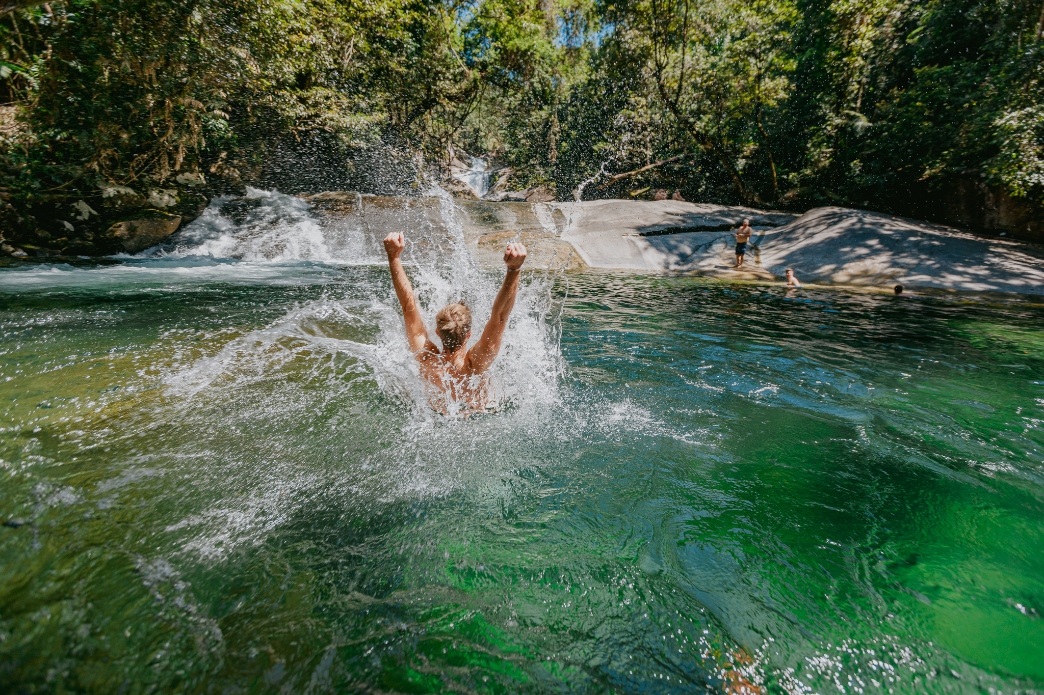 Adventurer joyfully splashes into crystal-clear green river beneath lush Daintree Rainforest canopy, with scenic waterfall behind.