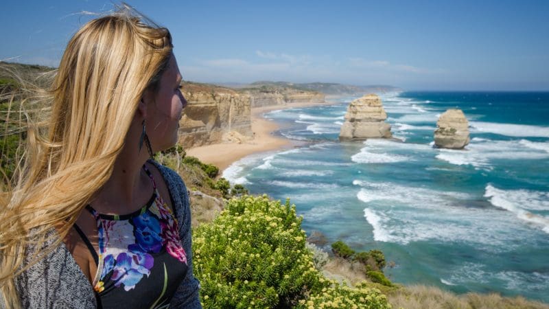 Blonde woman admires breathtaking ocean views on a sunny Great Ocean Road Reverse Eco Tour near iconic Twelve Apostles, Australia.