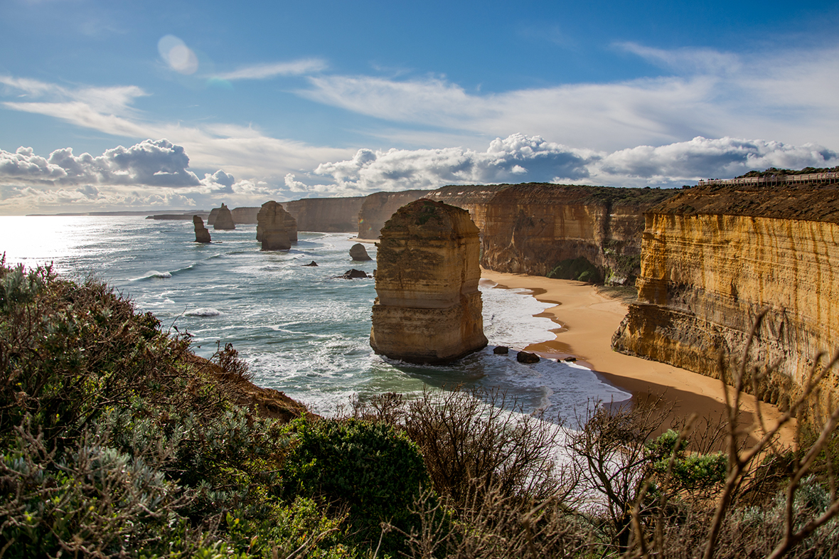 Majestic rock formations tower over sunlit coastline on the 1 Day Great Ocean Road Tour, framed by pristine beaches and rugged cliffs.