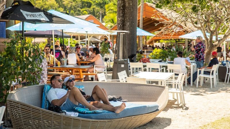 Couple lounging by the vibrant Falls To Paradise Waterfall and Oceanfront Beach Bar, surrounded by lush palm trees and tropical ambience.