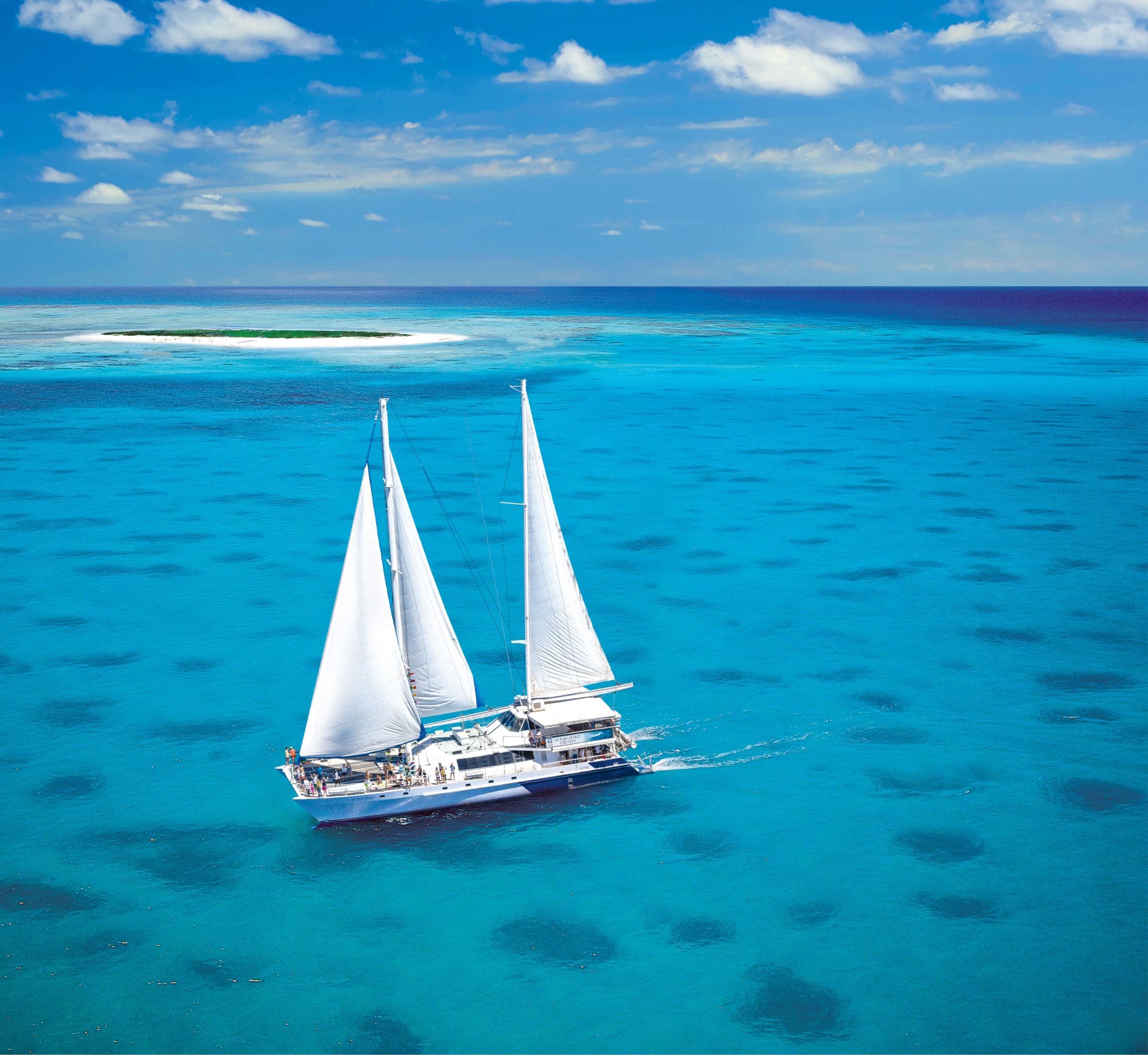 Ocean Spirit yacht with twin white sails cruising by Michaelmas Cay Reef under clear blue skies on a Great Barrier Reef tour.
