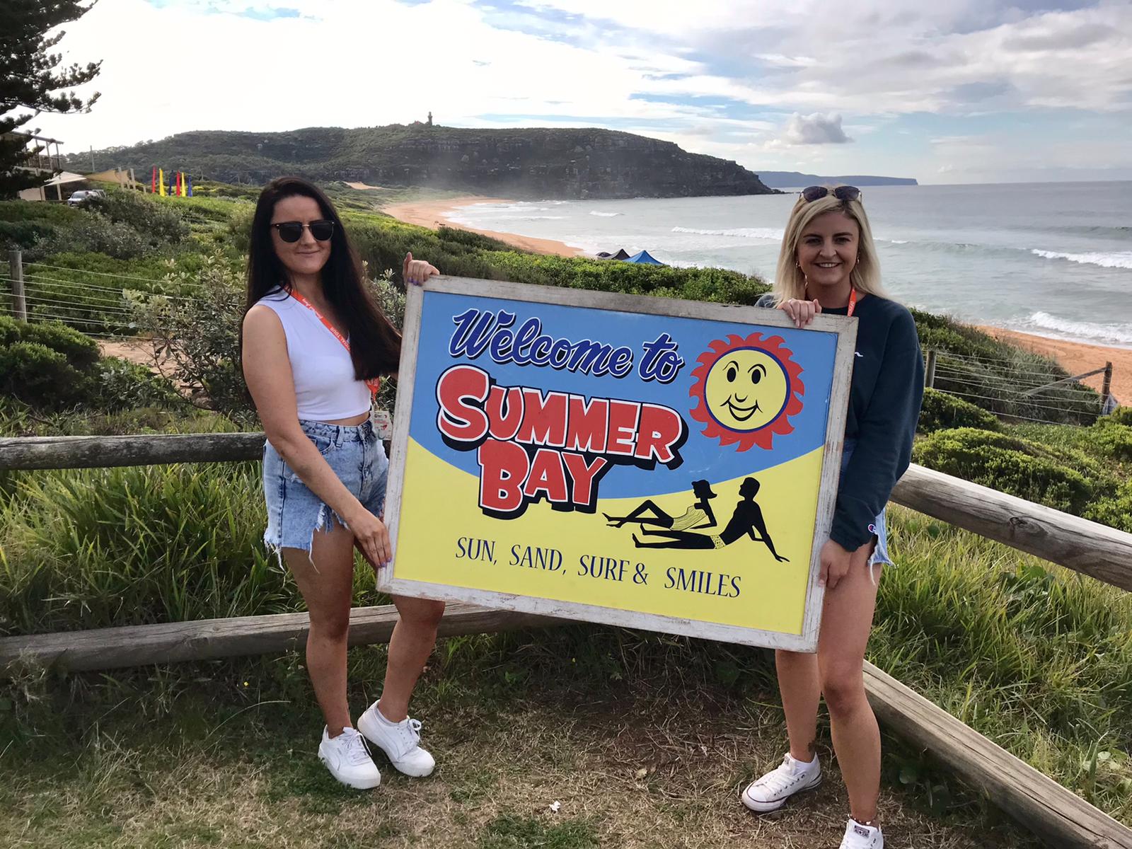 Two women on a sunlit Home Away Tour beach hold a “Welcome to Summer Bay” sign, with lush green hills and blue sea in view.