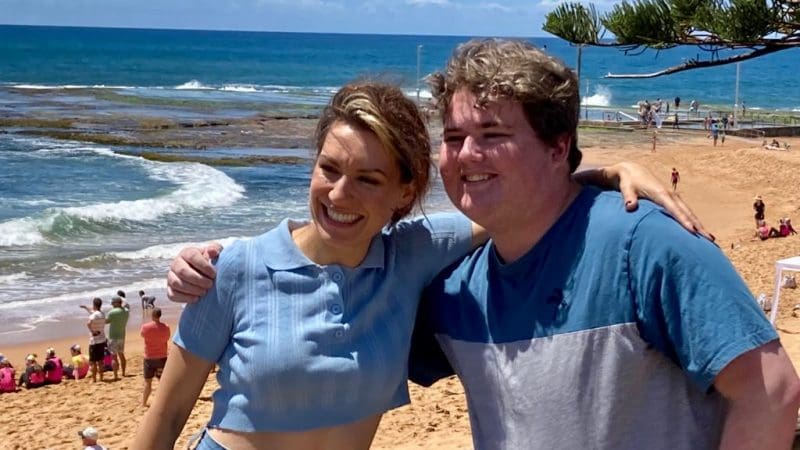 Happy travellers embrace and smile on a sunny beach during a 1 Day Home Away Tour, crystal blue ocean waves sparkling behind them.