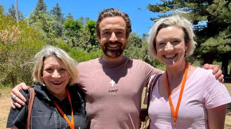 Three adults smiling outdoors wearing Celebtime Tours orange lanyards, enjoying a 1 Day Home Away Tour beneath a clear blue sky.