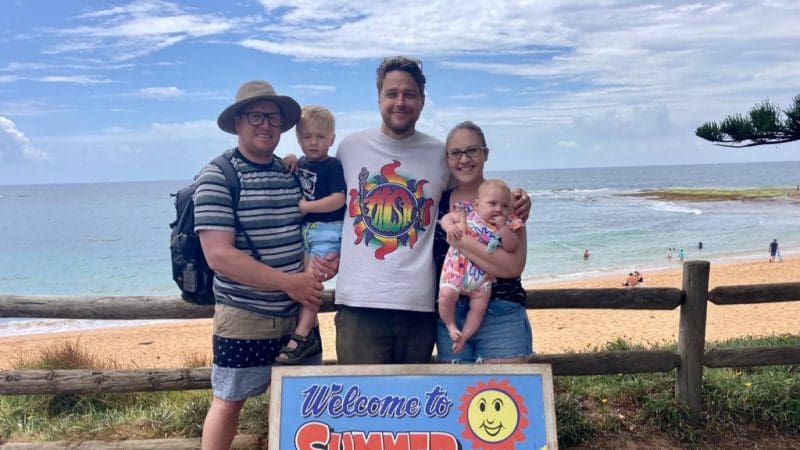 Group of three adults and two children smiling by a scenic beach, holding a Welcome to Summer sign during their 1 Day Home Away Tour.