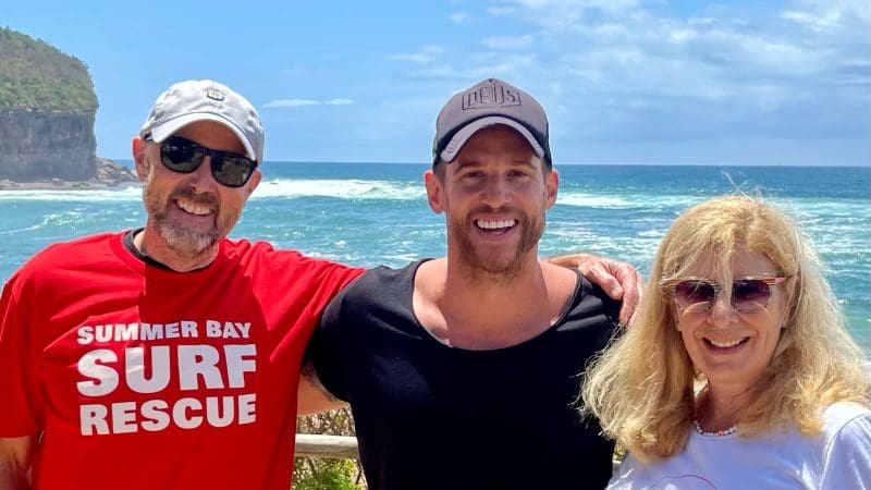 Three people smile by the sea in Summer Bay Surf Rescue shirts during a Celebtime Tours 1 Day Home and Away experience.
